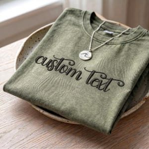May include: An olive green t-shirt with the embroidered text "custom text" in a cursive font. A silver necklace with a wave pendant rests on the shirt, which is folded in a ceramic bowl.
