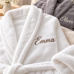 Puede incluir: Una bata de baño blanca de felpa con el nombre Emma bordado en letra cursiva marrón en el pecho y un cinturón a juego. Una segunda bata de baño gris oscuro con el nombre Oliver bordado en blanco es visible en el fondo.