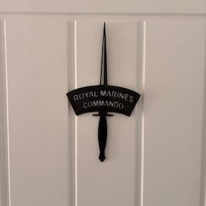 May include: Black metal Royal Marines Commando emblem. The emblem is in the shape of a dagger with a curved banner above the handle. The banner reads "ROYAL MARINES COMMANDO" in capital letters.