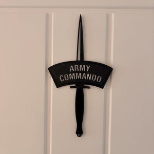 以下が含まれることがあります： 黒い金属製の壁の装飾で、白い文字で「ARMY COMMANDO」と書かれた短剣のデザインが特徴です。 短剣は湾曲した柄と長く尖った刃を持っています。 装飾は白いドアに取り付けられています。
