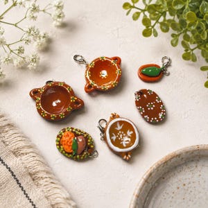 May include: A collection of six miniature clay charms shaped like traditional Mexican pottery, including small bowls, a plate with food, and a green chili, each featuring hand-painted details in brown, white, green, and yellow with metal clasps.