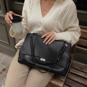 以下が含まれることがあります： シルバーカラーの長方形のツイストロック式留め具と2本の細いショルダーストラップが付いた黒いレザーのショルダーバッグ。バッグは質感のある仕上げで、クリーム色のケーブルニットのVネックセーターとベージュのズボンの前に配置されています。