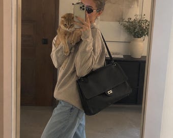 May include: A large black leather shoulder bag with a rectangular flap, a gold-toned turn-lock closure, and a long shoulder strap. The bag is worn over the shoulder, paired with a beige long-sleeved knit sweater and light blue denim jeans.