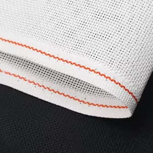 May include: Close-up of white mesh fabric with a red zigzag trim. The fabric is folded over, revealing the texture and the contrasting black background. The mesh has small, evenly spaced holes.
