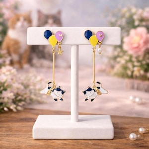 May include: A pair of gold-tone drop earrings featuring a cluster of three small enamel balloons in dark blue, yellow, and pink. A dangling gold star charm hangs from one earring, while a small white pearl bead hangs from the other, above a cat.