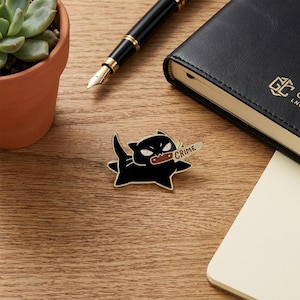 May include: A black enamel pin shaped like a cat with white eyes and a sharp expression, holding a small knife in its mouth. The blade of the knife features the word CRIME printed in black letters against a light-colored background.