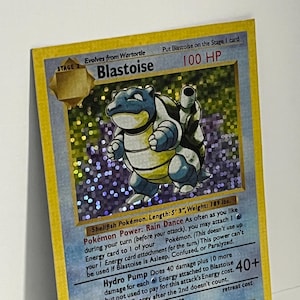 May include: A holographic Blastoise trading card featuring the blue Shellfish Pokémon with cannons on its back. The card displays 100 HP, the Rain Dance Pokémon Power, and the Hydro Pump attack. Text includes Length: 5' 3", Weight: 189 lbs.
