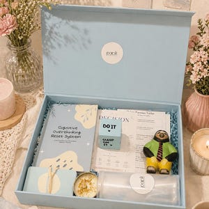 May include: A light blue box filled with self-care items. The box contains a book titled "Cognitive Overthinking Reset System", a small cube with "Do It" and "Sleep on it" printed on it, a candle, and a sloth figurine. The box has the brand name "panik" on it.