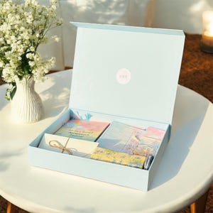 Könnte beinhalten: Eine offene, hellblaue Geschenkbox auf einem weißen Tisch, die Schreibwaren enthält. Darin befinden sich ein Journal, Notizkarten und andere Artikel. Links steht eine Vase mit weißen Blumen. Die Box hat einen kleinen, runden Aufkleber auf dem Deckel.