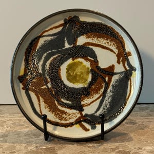 Può includere: Un piatto in ceramica con un design astratto unico. Il piatto presenta una base bianca con motivi a spirale marroni, neri e gialli. Il bordo è marrone scuro. Il piatto è esposto su un supporto in metallo nero.