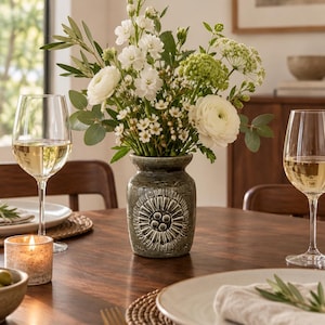 Op de afbeelding: Een kleine, donkergroene keramische vaas met een gestructureerd, cirkelvormig zonnepatroon aan de voorzijde, gevuld met een boeket van witte ranonkels, kleine witte bloemen, groen blad en olijftakken, geplaatst op een houten tafel.