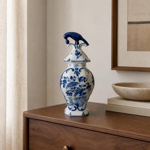 May include: A white ceramic vase with a hexagonal body and a matching lid featuring a dark blue bird-shaped finial. The vase is decorated with intricate blue floral and bird patterns painted in a traditional style across its entire surface.