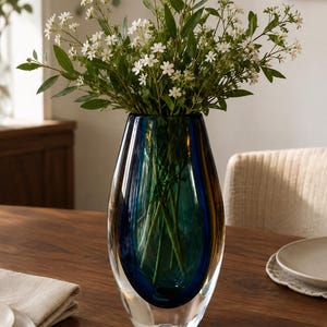 Könnte beinhalten: Eine hohe, tropfenförmige Glasvase mit einem tiefblauen und türkisfarbenen Farbverlauf im Inneren, umhüllt von klarem Glas. Die Vase enthält einen Strauß weißer Blumen mit grünen Blättern und trägt ein kleines Etikett mit der Aufschrift Kosta.