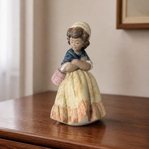 Può includere: Una statuetta in ceramica raffigurante una persona con una cuffia bianca, uno scialle blu scuro e una gonna lunga e testurizzata nei toni del giallo e del pesca. La figura ha capelli castani, braccia incrociate e porta un cestino rosa.