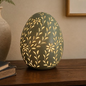 Op de afbeelding: Een eivormige saliegroene lamp met ingewikkelde uitgesneden patronen van bloemen en bladeren. Het licht schijnt vanuit de holle structuur en verlicht de bloemmotieven tegen het matte oppervlak van het keramiekachtige materiaal.