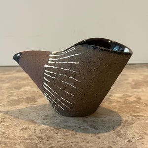May include: A handcrafted, dark brown ceramic bowl with a unique, asymmetrical shape. The bowl features a black rim and white linear accents on the exterior. The bowl is displayed on a marble-like surface.