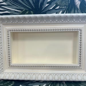 May include: A rectangular, off-white shadow box with ornate, raised detailing. The frame has a layered design with a recessed interior, perfect for displaying small items. The box is set against a patterned background.