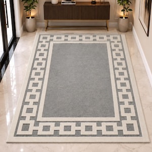 May include: A rectangular area rug featuring a solid gray center panel surrounded by a wide border with a repeating white geometric square pattern. The rug has a clean, flat-woven texture and is positioned on a light-colored tiled floor in a hallway.