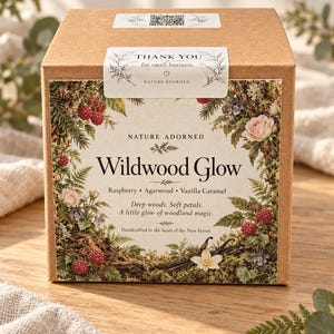 May include: A square brown cardboard box with a label featuring botanical illustrations of raspberries, flowers, and ferns. The text reads Nature Adorned, Wildwood Glow, Raspberry, Agarwood, Vanilla Caramel, Deep woods. Soft petals. A little glow of woodland magic. Handcrafted in the heart of the New Forest.