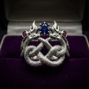 Op de afbeelding: Een zilveren ring met twee in elkaar verstrengelde slangen met gedetailleerde schubben. Een ronde, gefacetteerde blauwe edelsteen is aan de bovenkant tussen de slangenkoppen gezet, geflankeerd door twee kleinere, ronde, rode edelstenen.