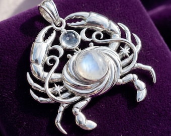May include: A silver crab pendant with a large, round, iridescent stone at its center and a smaller, clear stone above. The pendant has detailed claws, legs, and decorative swirls, set against a deep purple background.