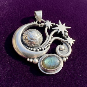May include: A silver pendant featuring a crescent moon, a textured sphere, and three star shapes. The design includes a bezel-set oval labradorite gemstone with blue and green iridescence, accented by small silver spheres and swirling metal details.
