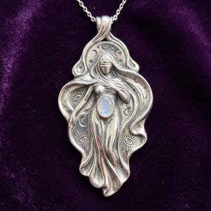 May include: A silver pendant necklace featuring a blindfolded figure with flowing hair and garments. The design includes celestial motifs like moons, stars, and eyes, with a central oval moonstone cabochon set into the figure's abdomen.