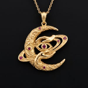 May include: A gold-colored pendant necklace featuring a crescent moon design with ornate scrollwork and small red gemstones. The pendant includes an eye and hands, suspended from a delicate chain.