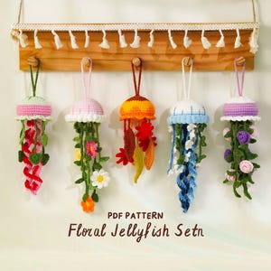 Puede incluir: Cinco adornos de medusas de ganchillo hechas a mano en varios colores cuelgan de un estante de madera. Cada medusa presenta una combinación de colores diferente, incluyendo rosa, naranja, azul y morado, con zarcillos y flores decorativas. El texto "PDF PATTERN Floral Jellyfish Setn" es visible.