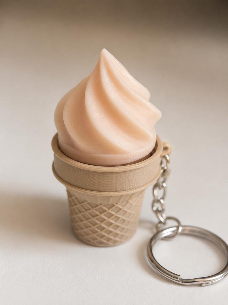 May include: A keychain featuring a light beige ice cream cone with a swirled, pale pink soft-serve top. The cone has a textured waffle pattern and is attached to a silver-toned metal chain and a circular split ring for keys.