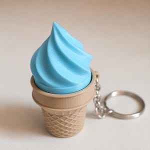 May include: A keychain featuring a light blue soft-serve ice cream swirl set inside a tan, textured waffle cone. The ice cream is shaped with a pointed top and spiral ridges. A silver-toned metal chain and circular ring are attached to the side.