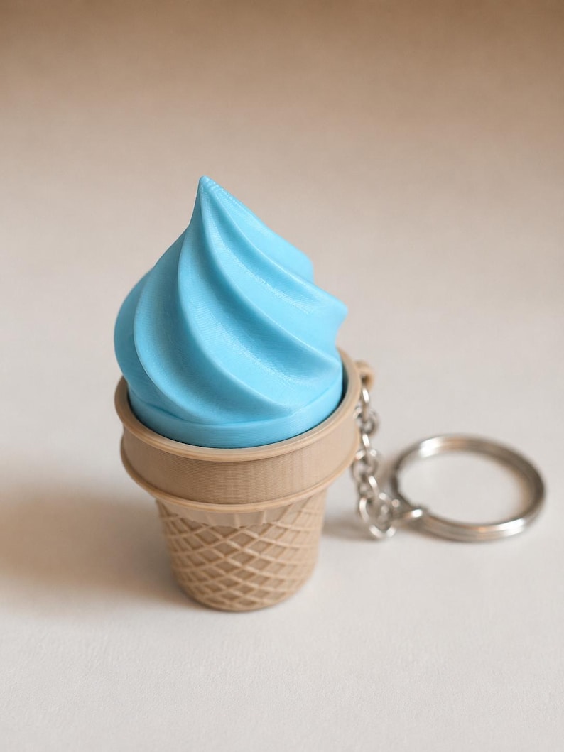 May include: A keychain featuring a light blue soft-serve ice cream swirl set inside a tan, textured waffle cone. The ice cream is shaped with a pointed top and spiral ridges. A silver-toned metal chain and circular ring are attached to the side.