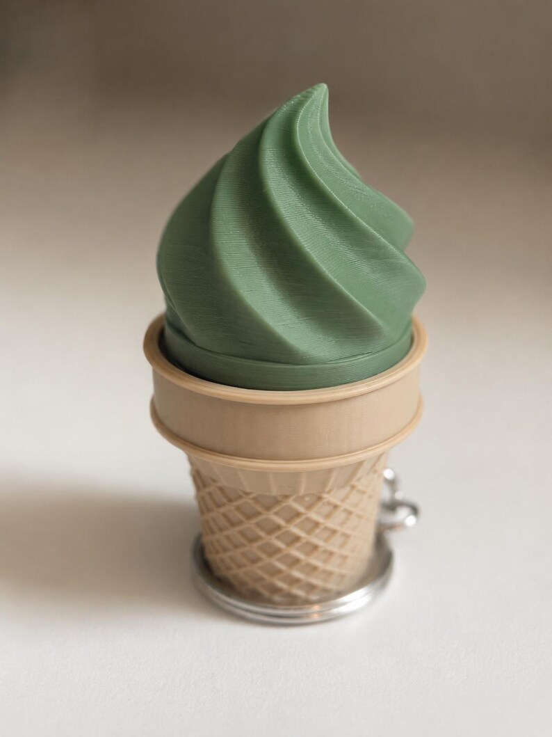 May include: A keychain featuring a sage green, swirled soft-serve ice cream top set inside a light tan, textured waffle cone base. The object is constructed with a visible 3D printed layered finish and includes a silver metal ring attachment.