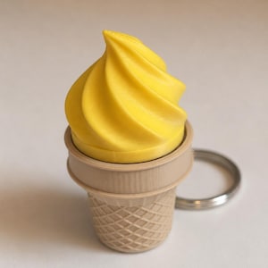 May include: A keychain featuring a bright yellow soft-serve ice cream swirl sitting inside a light tan waffle cone base. The cone has a textured, cross-hatched grid pattern on its exterior and is attached to a circular silver metal ring.