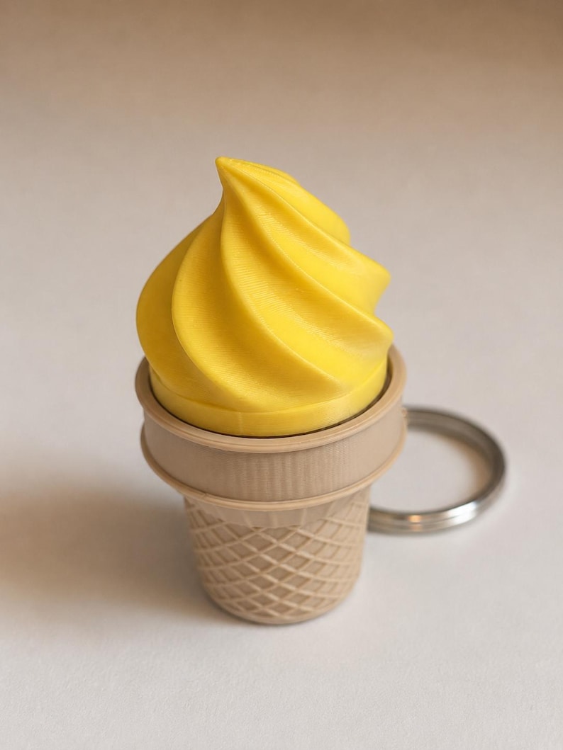 May include: A keychain featuring a bright yellow soft-serve ice cream swirl sitting inside a light tan waffle cone base. The cone has a textured, cross-hatched grid pattern on its exterior and is attached to a circular silver metal ring.