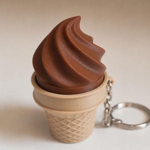 May include: A chocolate soft serve ice cream cone keychain featuring a brown swirled top and a light tan waffle cone base with a textured grid pattern. A silver metal chain and a circular split ring are attached to the side of the cone.