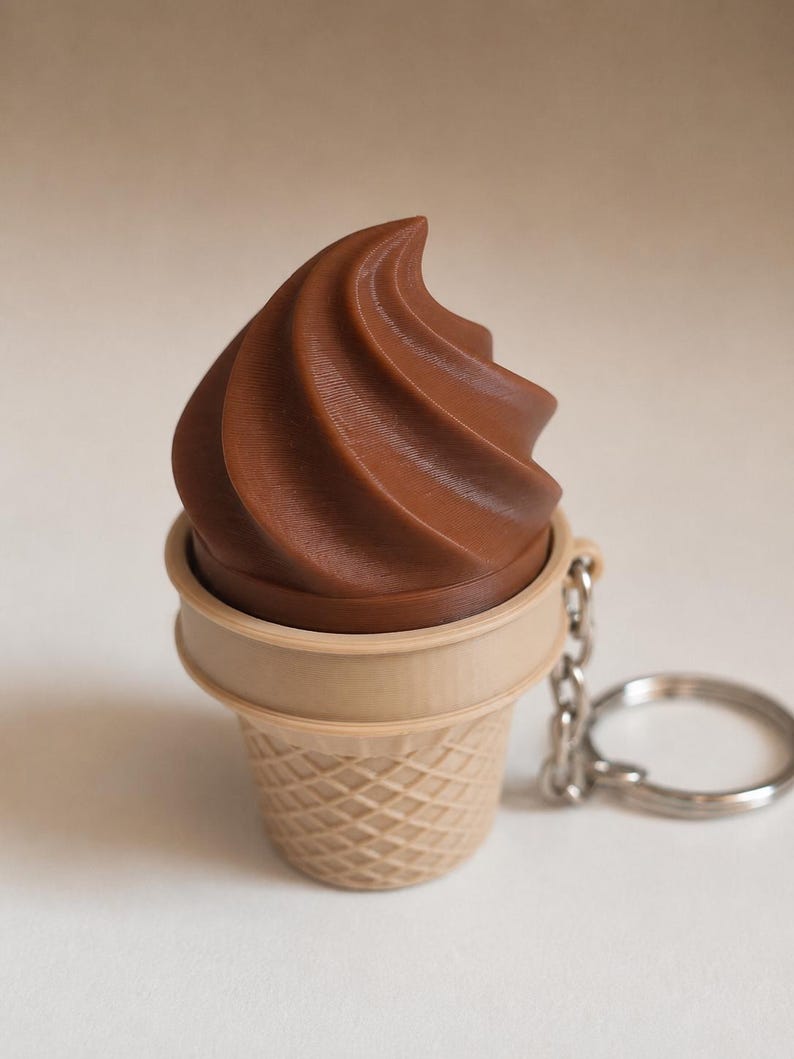 May include: A chocolate soft serve ice cream cone keychain featuring a brown swirled top and a light tan waffle cone base with a textured grid pattern. A silver metal chain and a circular split ring are attached to the side of the cone.
