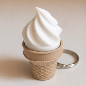 May include: A keychain featuring a white, swirled soft-serve ice cream top set inside a light brown, textured waffle cone base. The object includes a circular silver metal ring attached to the side for securing keys or other small items.