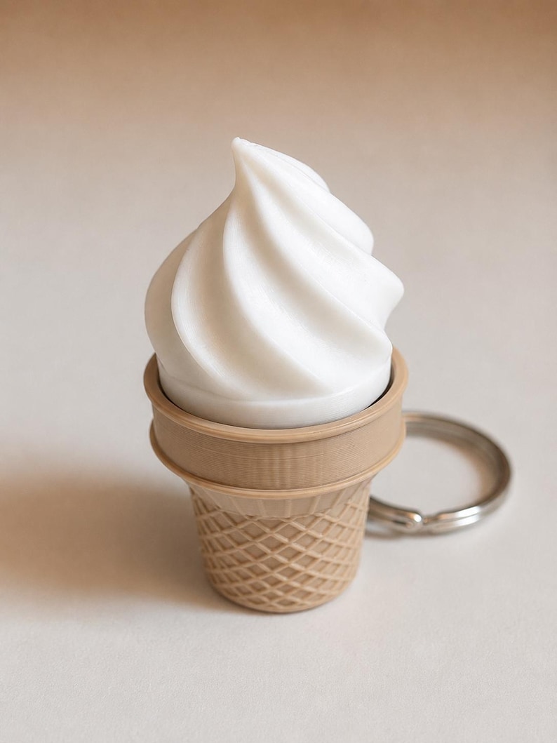 May include: A keychain featuring a white, swirled soft-serve ice cream top set inside a light brown, textured waffle cone base. The object includes a circular silver metal ring attached to the side for securing keys or other small items.