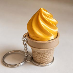 May include: A keychain featuring a shiny golden swirl of soft-serve ice cream sitting inside a light tan, textured waffle cone. The ice cream and cone are attached to a standard silver-colored metal split ring by a short silver metal chain.