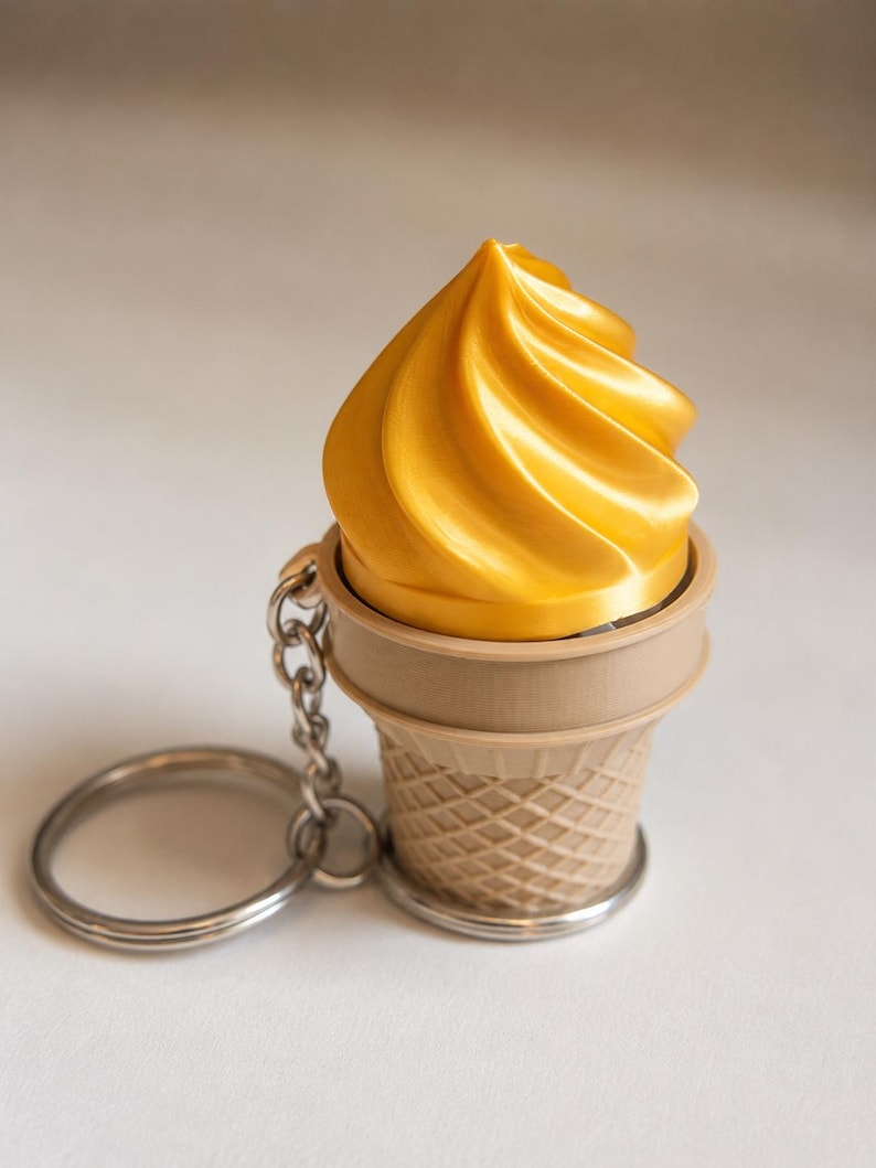May include: A keychain featuring a shiny golden swirl of soft-serve ice cream sitting inside a light tan, textured waffle cone. The ice cream and cone are attached to a standard silver-colored metal split ring by a short silver metal chain.