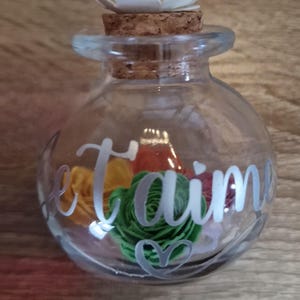 Puede incluir: Un tarro de cristal transparente con un tapón de corcho y una flor blanca en la parte superior. El tarro contiene flores de colores y las palabras "Je t'aime" están escritas en el lateral. El tarro está sobre una superficie de madera.