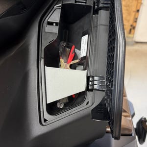 May include: A black vehicle compartment with a storage area containing keys, a white card, and other items. The compartment door is open, revealing a honeycomb-patterned grill.