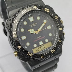 May include: A black Casio Quartz analog-digital watch with a rotating bezel featuring white numerals. The black dial displays luminous hour markers, gold-toned hands, and the text Casio Quartz 100M Alarm Chronograph. A digital display shows 12:00.