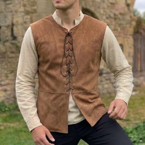 May include: A brown suede vest with a lace-up front. The vest is sleeveless and has a high collar. The vest is worn over a long-sleeved beige shirt. The background is a stone wall.