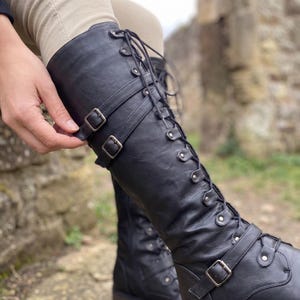 May include: A black knee-high boot featuring a lace-up front with silver-toned eyelets and two horizontal straps across the upper shaft, each secured with a rectangular metal buckle. The boot has a smooth finish and a structured, tall silhouette.