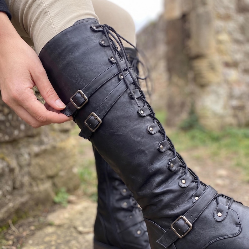 May include: A black knee-high boot featuring a lace-up front with silver-toned eyelets and two horizontal straps across the upper shaft, each secured with a rectangular metal buckle. The boot has a smooth finish and a structured, tall silhouette.