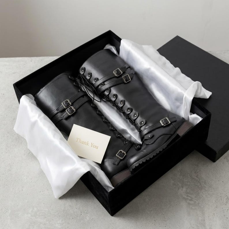 May include: A pair of black knee-high boots with lace-up fronts and multiple adjustable buckle straps, presented in a black gift box lined with white tissue paper. A small white card with the printed text Thank You rests on the boots.