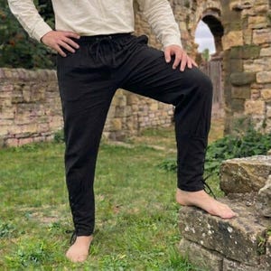 Puede incluir: Pantalones medievales negros con cintura con cordón y detalles de cordones en los tobillos. Los pantalones están hechos de una tela ligera y son adecuados para recreaciones históricas o eventos de disfraces. Los pantalones se muestran al aire libre sobre hierba.