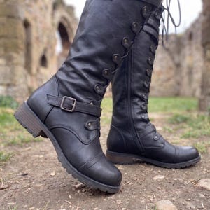 May include: A pair of black mid-calf boots featuring a lace-up front with metal eyelets, a side zipper closure, and a decorative strap with a silver-toned buckle across the ankle. The boots have a rounded toe, a low block heel, and a textured sole.